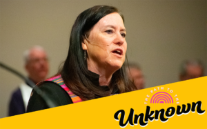 A woman pastor preaching in a black robe with a brightly multi-colored stole.